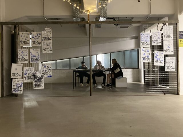 Drei junge Leute sitzen in einer ehemaligen Garagen-Nische im Stilwerk in den Kant-Garagen. Rechts und links der Nische hängen Plakate zur Werkschau 2023.
