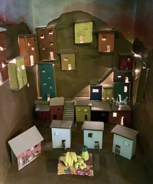 Einige kleine Hütten an einem Berghang. Im Zentrum ein Tisch mit Bananen.